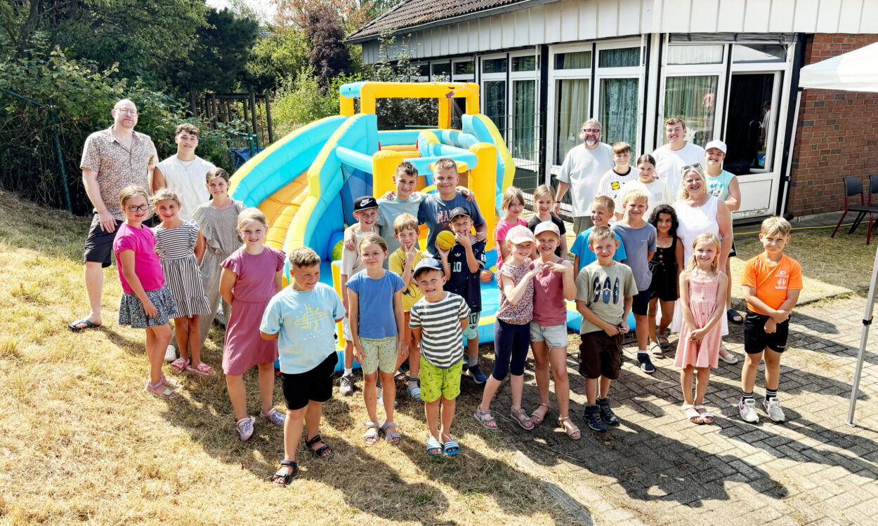 Eine große Gruppe von Kindern und mehreren Erwachsenen steht bei sonnigem Wetter vor einer bunt gelb-blauen Hüpfburg. Die Kinder lächeln in die Kamera und tragen sommerliche Kleidung. Im Hintergrund befindet sich ein Gebäude mit großen Fenstern, umgeben von Bäumen und Sträuchern.