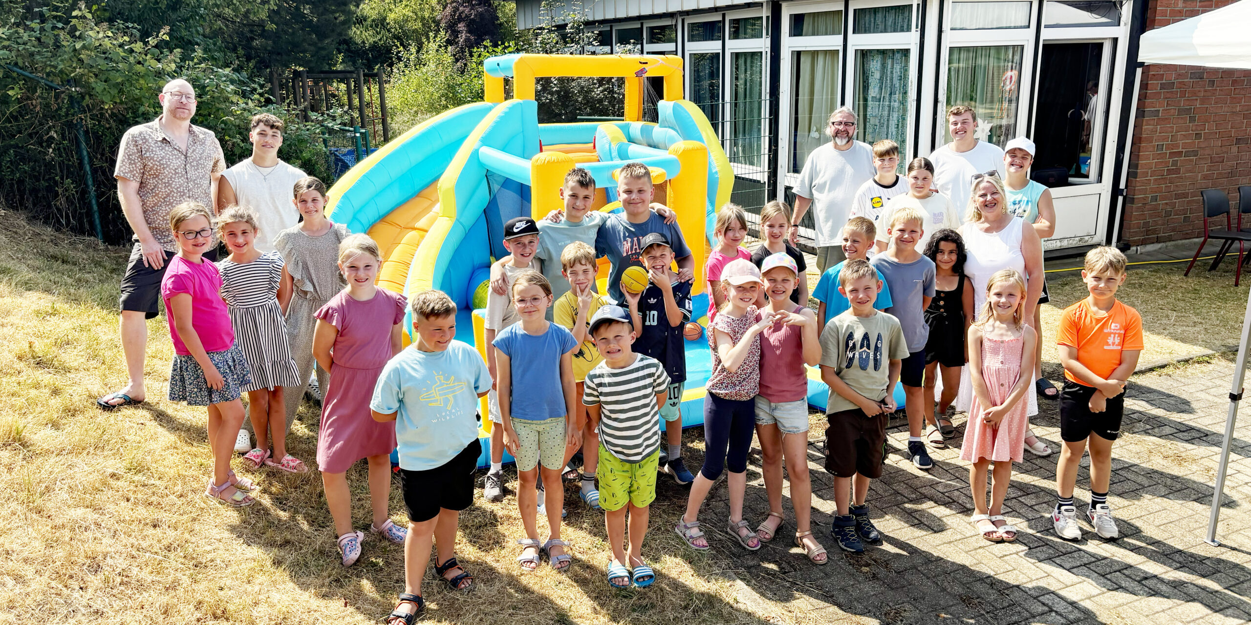 Eine große Gruppe von Kindern und mehreren Erwachsenen steht bei sonnigem Wetter vor einer bunt gelb-blauen Hüpfburg. Die Kinder lächeln in die Kamera und tragen sommerliche Kleidung. Im Hintergrund befindet sich ein Gebäude mit großen Fenstern, umgeben von Bäumen und Sträuchern.