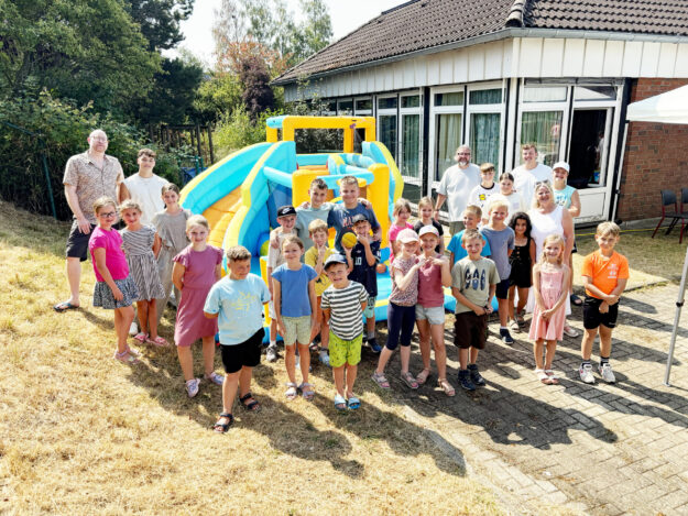 Eine große Gruppe von Kindern und mehreren Erwachsenen steht bei sonnigem Wetter vor einer bunt gelb-blauen Hüpfburg. Die Kinder lächeln in die Kamera und tragen sommerliche Kleidung. Im Hintergrund befindet sich ein Gebäude mit großen Fenstern, umgeben von Bäumen und Sträuchern.