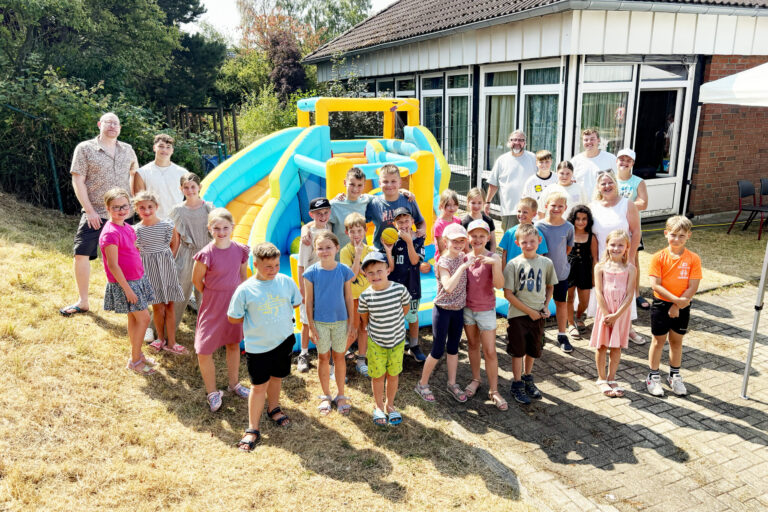 Foto: Stadt Altena Eine große Gruppe von Kindern und mehreren Erwachsenen steht bei sonnigem Wetter vor einer bunt gelb-blauen Hüpfburg. Die Kinder lächeln in die Kamera und tragen sommerliche Kleidung. Im Hintergrund befindet sich ein Gebäude mit großen Fenstern, umgeben von Bäumen und Sträuchern.