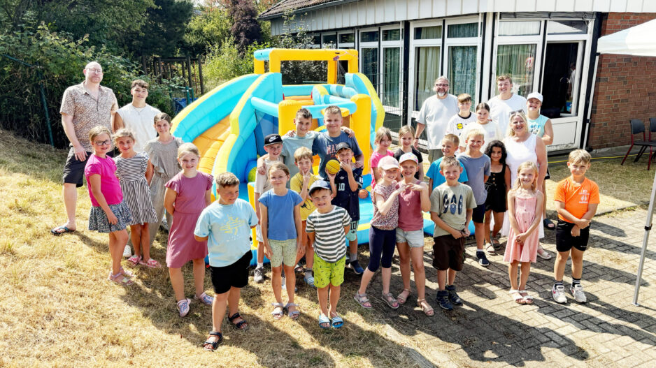 Eine große Gruppe von Kindern und mehreren Erwachsenen steht bei sonnigem Wetter vor einer bunt gelb-blauen Hüpfburg. Die Kinder lächeln in die Kamera und tragen sommerliche Kleidung. Im Hintergrund befindet sich ein Gebäude mit großen Fenstern, umgeben von Bäumen und Sträuchern.