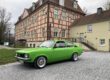 Ein leuchtend grüner Oldtimer steht auf einem hellen Kiesplatz vor einem historischen Fachwerkgebäude mit rosafarbener Fassade und dunklen Holzbalken. Der restaurierte Zweitürer ist seitlich schräg zur Kamera ausgerichtet und hebt sich deutlich von der ruhigen Architektur im Hintergrund ab. Die Aufnahme vermittelt den Eindruck einer Oldtimerpräsentation vor historischer Kulisse.