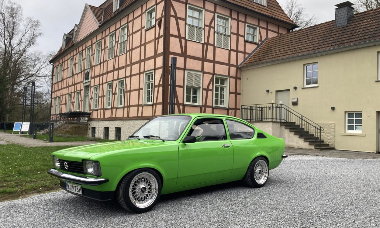Ein leuchtend grüner Oldtimer steht auf einem hellen Kiesplatz vor einem historischen Fachwerkgebäude mit rosafarbener Fassade und dunklen Holzbalken. Der restaurierte Zweitürer ist seitlich schräg zur Kamera ausgerichtet und hebt sich deutlich von der ruhigen Architektur im Hintergrund ab. Die Aufnahme vermittelt den Eindruck einer Oldtimerpräsentation vor historischer Kulisse.