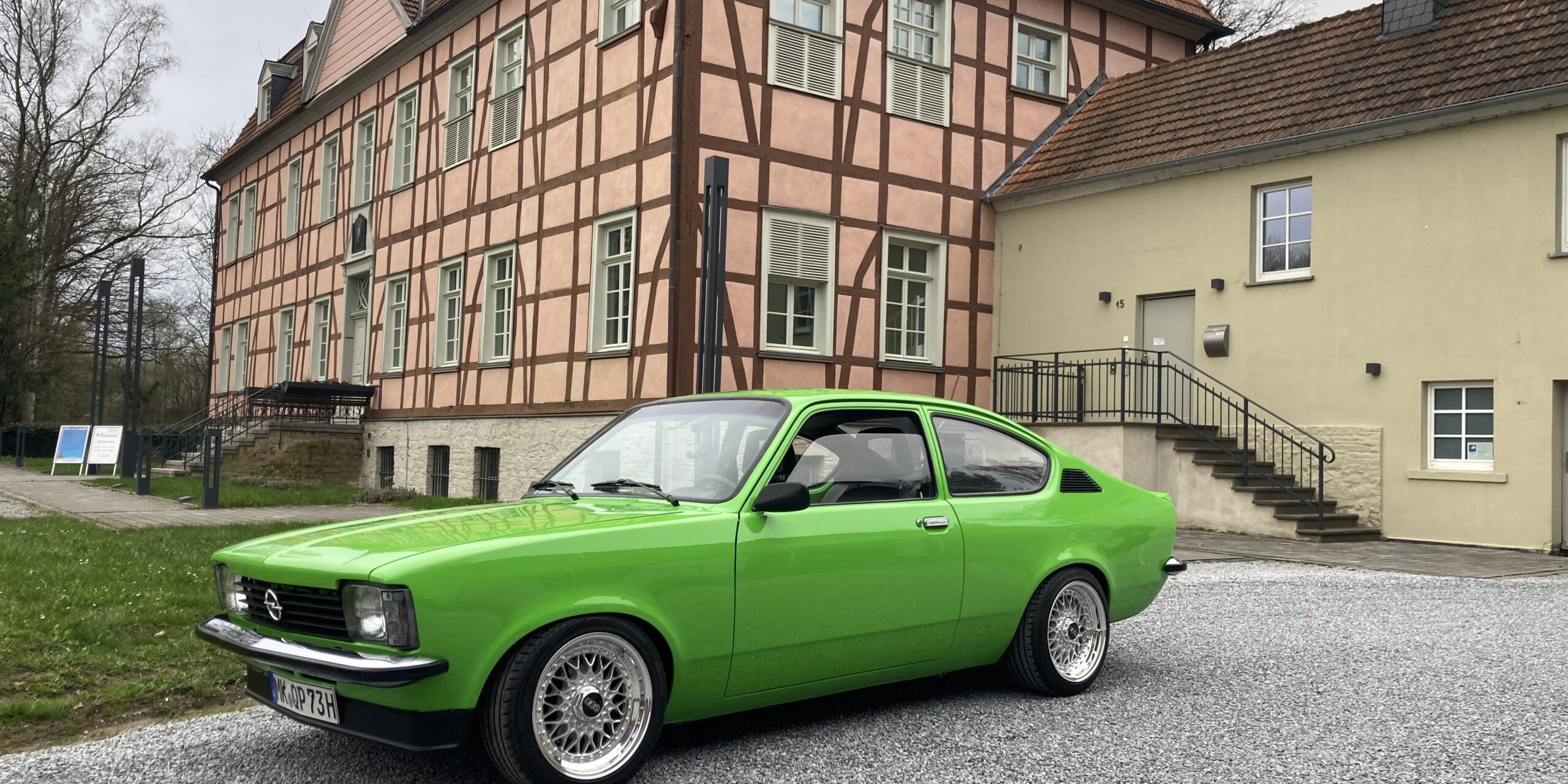 Ein leuchtend grüner Oldtimer steht auf einem hellen Kiesplatz vor einem historischen Fachwerkgebäude mit rosafarbener Fassade und dunklen Holzbalken. Der restaurierte Zweitürer ist seitlich schräg zur Kamera ausgerichtet und hebt sich deutlich von der ruhigen Architektur im Hintergrund ab. Die Aufnahme vermittelt den Eindruck einer Oldtimerpräsentation vor historischer Kulisse.