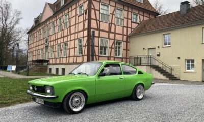 Ein leuchtend grüner Oldtimer steht auf einem hellen Kiesplatz vor einem historischen Fachwerkgebäude mit rosafarbener Fassade und dunklen Holzbalken. Der restaurierte Zweitürer ist seitlich schräg zur Kamera ausgerichtet und hebt sich deutlich von der ruhigen Architektur im Hintergrund ab. Die Aufnahme vermittelt den Eindruck einer Oldtimerpräsentation vor historischer Kulisse.
