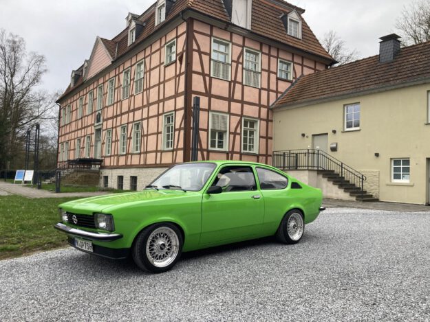 Foto: Sascha Krause Ein leuchtend grüner Oldtimer steht auf einem hellen Kiesplatz vor einem historischen Fachwerkgebäude mit rosafarbener Fassade und dunklen Holzbalken. Der restaurierte Zweitürer ist seitlich schräg zur Kamera ausgerichtet und hebt sich deutlich von der ruhigen Architektur im Hintergrund ab. Die Aufnahme vermittelt den Eindruck einer Oldtimerpräsentation vor historischer Kulisse.