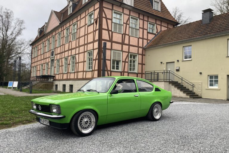 Ein leuchtend grüner Oldtimer steht auf einem hellen Kiesplatz vor einem historischen Fachwerkgebäude mit rosafarbener Fassade und dunklen Holzbalken. Der restaurierte Zweitürer ist seitlich schräg zur Kamera ausgerichtet und hebt sich deutlich von der ruhigen Architektur im Hintergrund ab. Die Aufnahme vermittelt den Eindruck einer Oldtimerpräsentation vor historischer Kulisse.