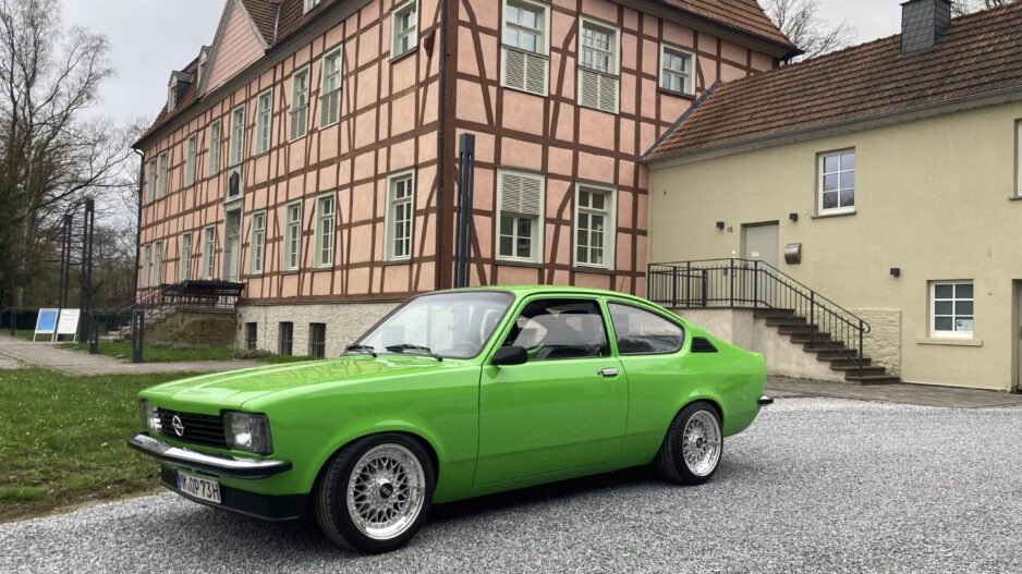 Ein leuchtend grüner Oldtimer steht auf einem hellen Kiesplatz vor einem historischen Fachwerkgebäude mit rosafarbener Fassade und dunklen Holzbalken. Der restaurierte Zweitürer ist seitlich schräg zur Kamera ausgerichtet und hebt sich deutlich von der ruhigen Architektur im Hintergrund ab. Die Aufnahme vermittelt den Eindruck einer Oldtimerpräsentation vor historischer Kulisse.