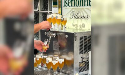 Eine Person zapft helles Bier aus einer Schankanlage in ein Glas. Rund um die Anlage stehen zahlreiche gefüllte Biergläser; im Hintergrund ist ein Schild mit der Aufschrift „Iserlohner Pilsener“ zu sehen.