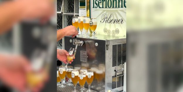 Eine Person zapft helles Bier aus einer Schankanlage in ein Glas. Rund um die Anlage stehen zahlreiche gefüllte Biergläser; im Hintergrund ist ein Schild mit der Aufschrift „Iserlohner Pilsener“ zu sehen.