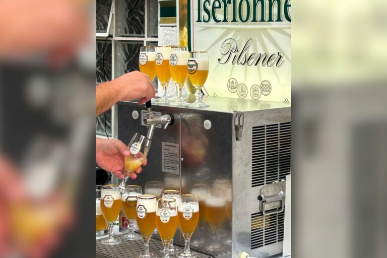 Eine Person zapft helles Bier aus einer Schankanlage in ein Glas. Rund um die Anlage stehen zahlreiche gefüllte Biergläser; im Hintergrund ist ein Schild mit der Aufschrift „Iserlohner Pilsener“ zu sehen.
