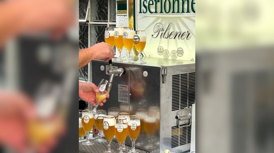 Eine Person zapft helles Bier aus einer Schankanlage in ein Glas. Rund um die Anlage stehen zahlreiche gefüllte Biergläser; im Hintergrund ist ein Schild mit der Aufschrift „Iserlohner Pilsener“ zu sehen.