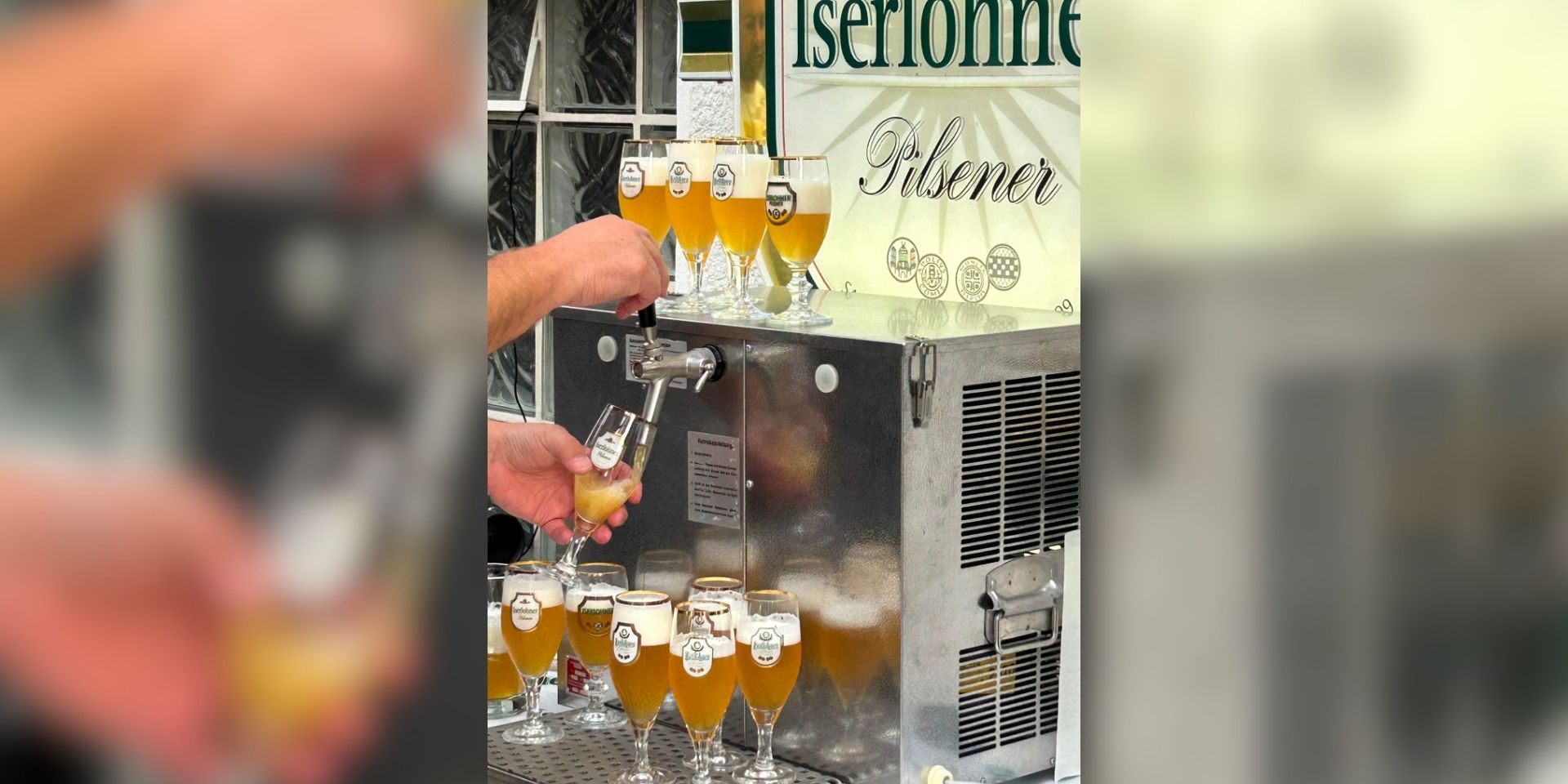 Eine Person zapft helles Bier aus einer Schankanlage in ein Glas. Rund um die Anlage stehen zahlreiche gefüllte Biergläser; im Hintergrund ist ein Schild mit der Aufschrift „Iserlohner Pilsener“ zu sehen.