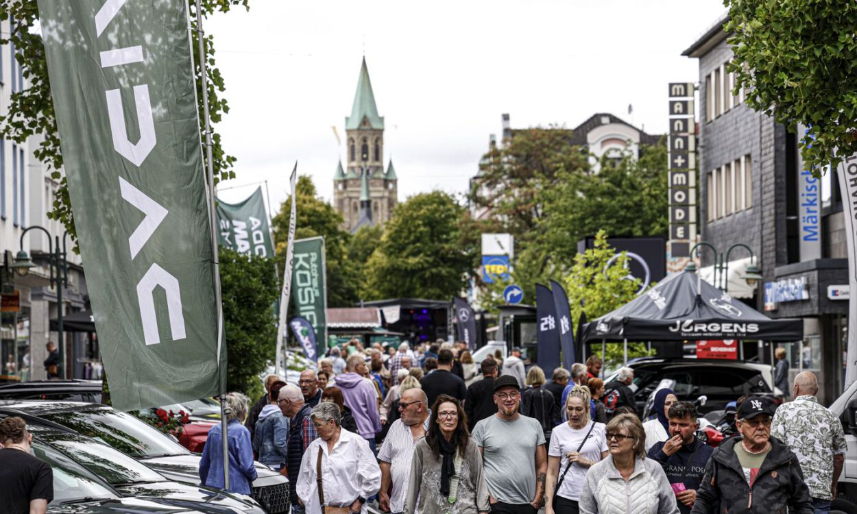 Eine belebte Einkaufsstraße während einer Veranstaltung. Zahlreiche Menschen gehen zwischen Verkaufsständen, Werbefahnen und ausgestellten Fahrzeugen entlang. Im Hintergrund ragt ein Kirchturm über die von Bäumen gesäumte Innenstadt.