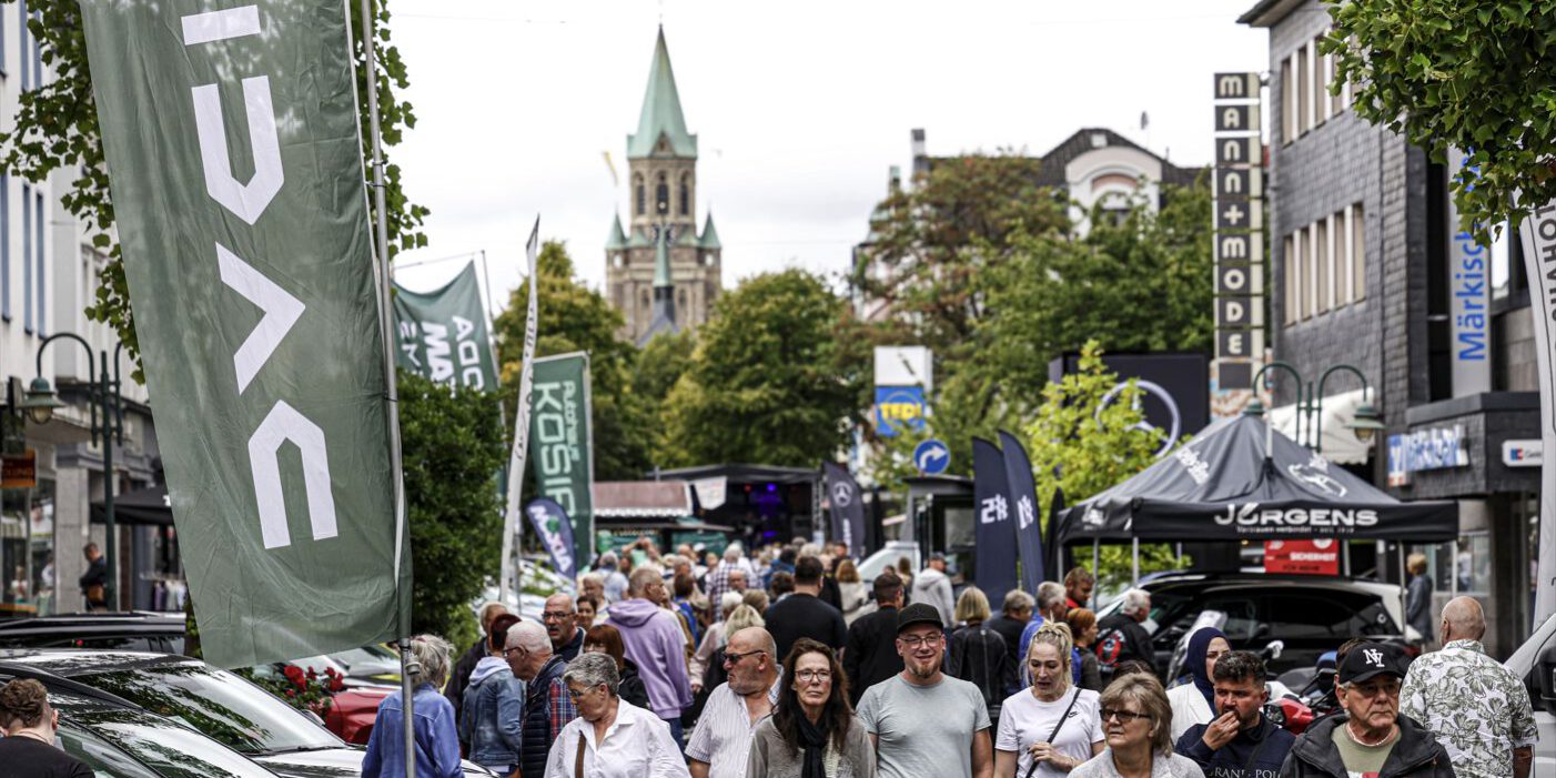 Eine belebte Einkaufsstraße während einer Veranstaltung. Zahlreiche Menschen gehen zwischen Verkaufsständen, Werbefahnen und ausgestellten Fahrzeugen entlang. Im Hintergrund ragt ein Kirchturm über die von Bäumen gesäumte Innenstadt.