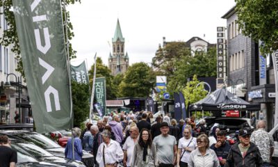 Eine belebte Einkaufsstraße während einer Veranstaltung. Zahlreiche Menschen gehen zwischen Verkaufsständen, Werbefahnen und ausgestellten Fahrzeugen entlang. Im Hintergrund ragt ein Kirchturm über die von Bäumen gesäumte Innenstadt.