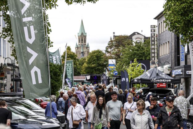 Foto: Dennis Echtermann Eine belebte Einkaufsstraße während einer Veranstaltung. Zahlreiche Menschen gehen zwischen Verkaufsständen, Werbefahnen und ausgestellten Fahrzeugen entlang. Im Hintergrund ragt ein Kirchturm über die von Bäumen gesäumte Innenstadt.