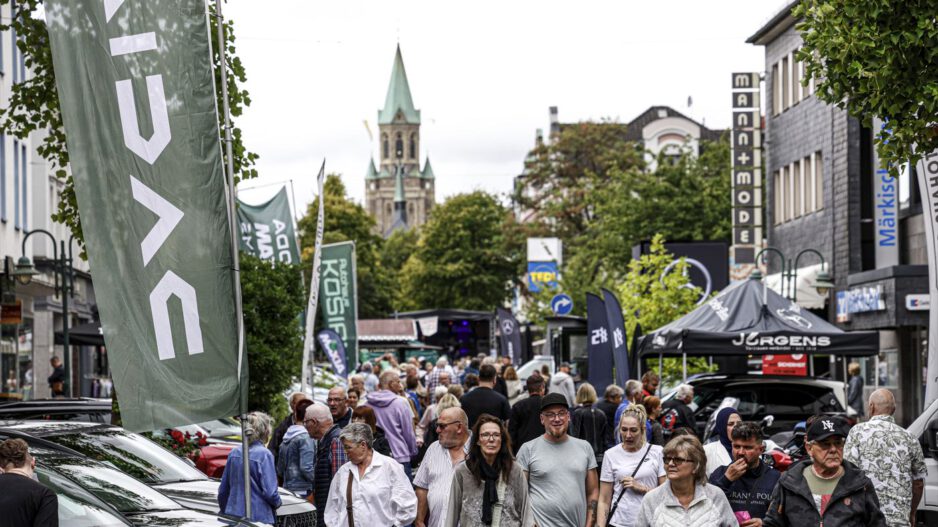 Eine belebte Einkaufsstraße während einer Veranstaltung. Zahlreiche Menschen gehen zwischen Verkaufsständen, Werbefahnen und ausgestellten Fahrzeugen entlang. Im Hintergrund ragt ein Kirchturm über die von Bäumen gesäumte Innenstadt.