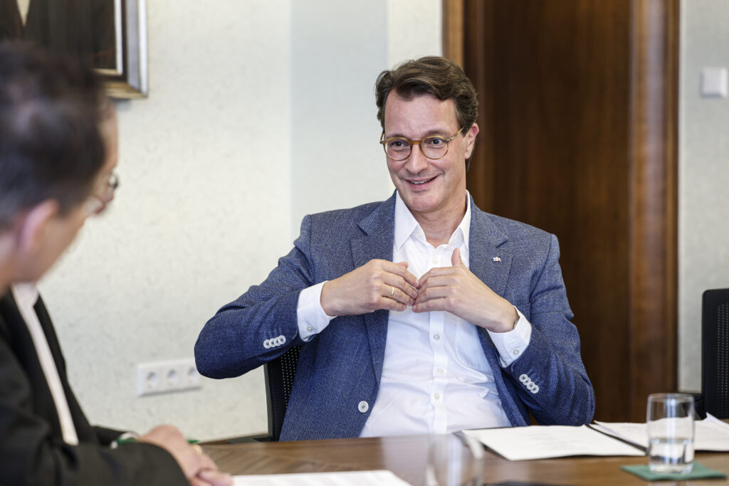 Hendrik Wüst sitzt mit Brille und blauem Sakko an einem Besprechungstisch und spricht lächelnd mit einer ihm gegenübersitzenden Person. Vor ihnen liegen Unterlagen und ein Wasserglas auf dem Tisch.