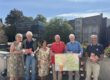 Sechs Personen stehen bei sonnigem Wetter auf einer Terrasse und präsentieren Wanderkarten sowie Informationsbroschüren. Zwei Männer halten eine großformatige Karte der Umgebung, während die anderen Prospekte in den Händen haben. Im Hintergrund sind Bäume, Blumenkästen und Gebäude der Innenstadt zu sehen.