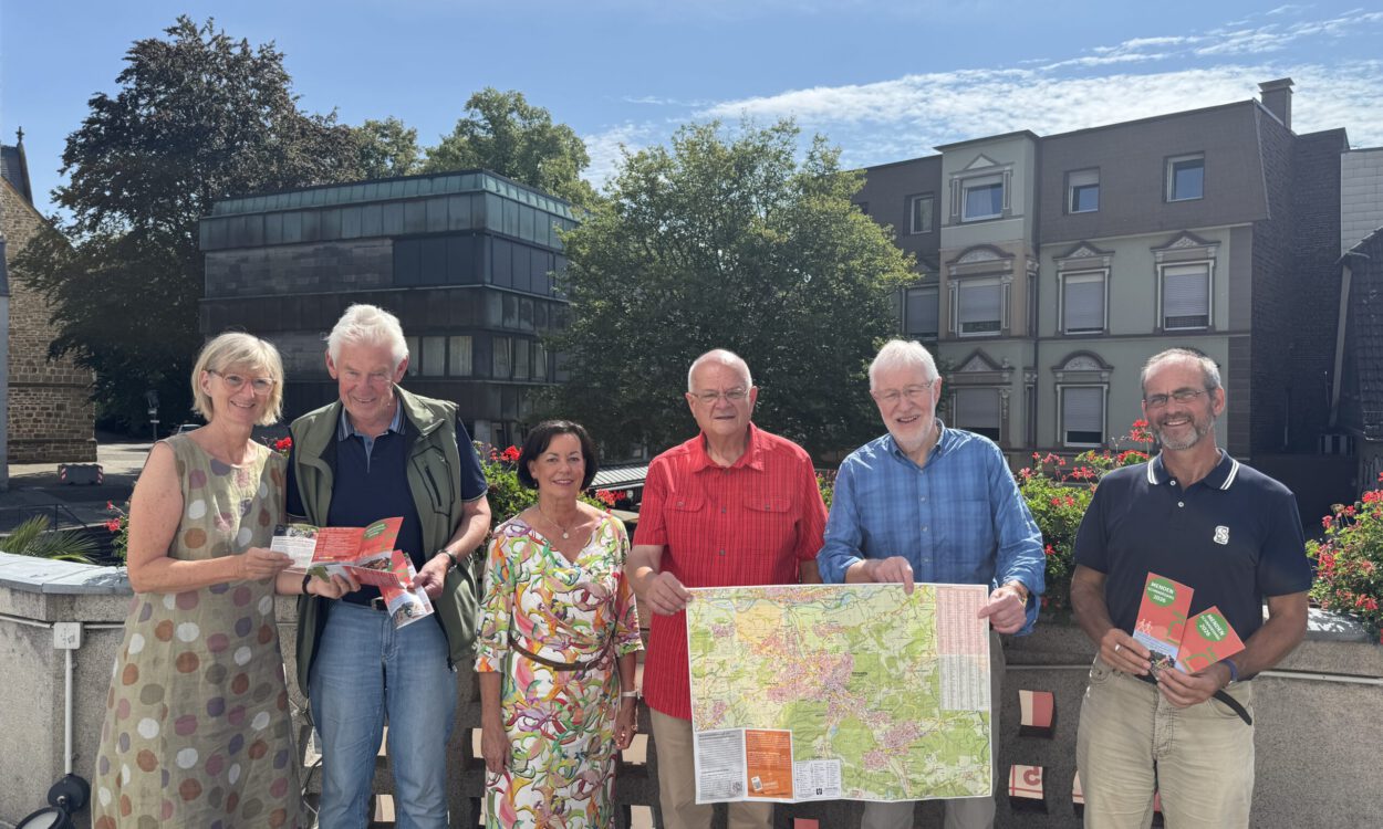 Sechs Personen stehen bei sonnigem Wetter auf einer Terrasse und präsentieren Wanderkarten sowie Informationsbroschüren. Zwei Männer halten eine großformatige Karte der Umgebung, während die anderen Prospekte in den Händen haben. Im Hintergrund sind Bäume, Blumenkästen und Gebäude der Innenstadt zu sehen.
