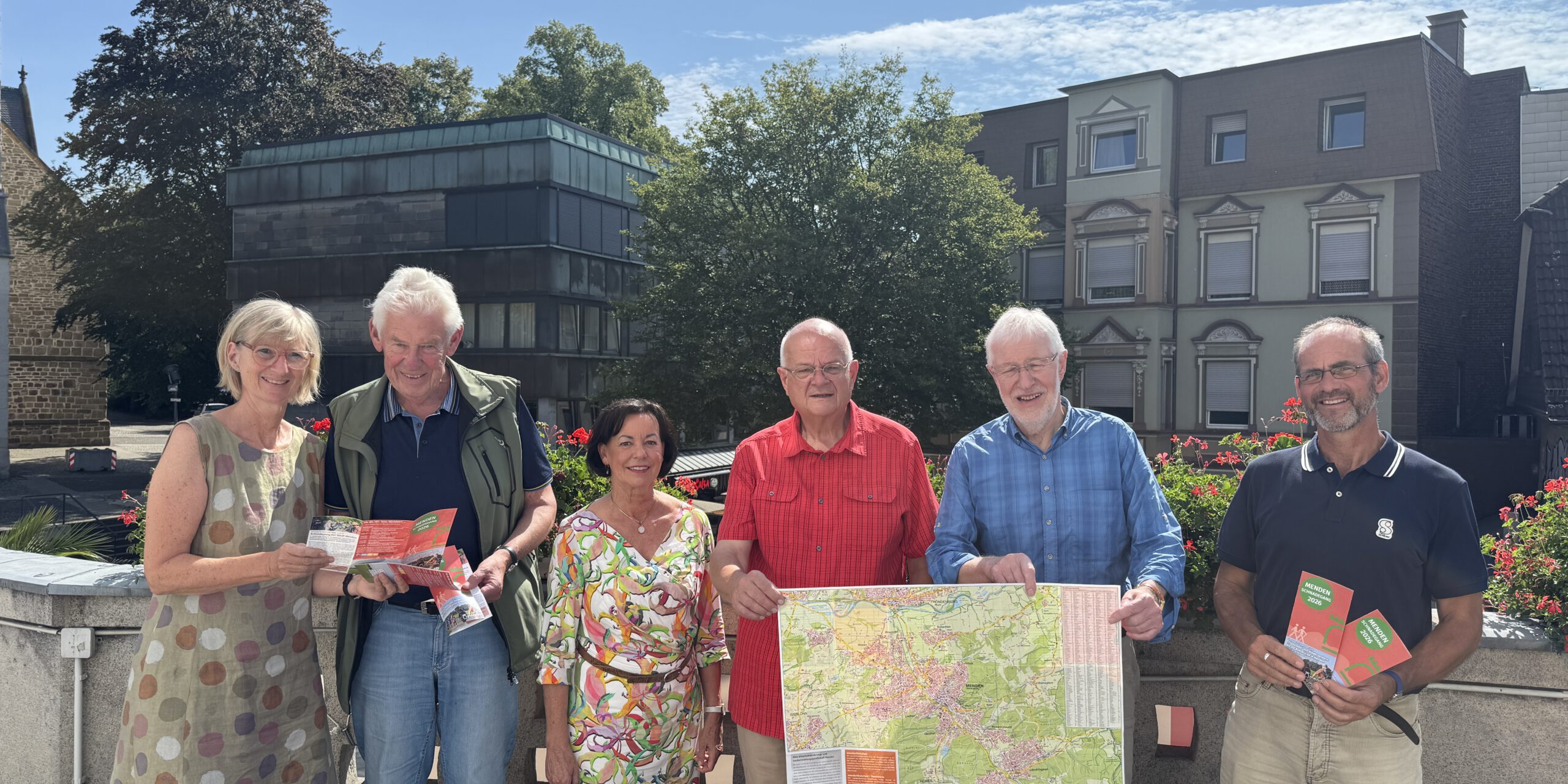 Sechs Personen stehen bei sonnigem Wetter auf einer Terrasse und präsentieren Wanderkarten sowie Informationsbroschüren. Zwei Männer halten eine großformatige Karte der Umgebung, während die anderen Prospekte in den Händen haben. Im Hintergrund sind Bäume, Blumenkästen und Gebäude der Innenstadt zu sehen.