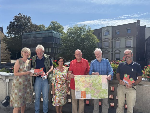 Foto: Denise Rüscher/Stadt Menden Sechs Personen stehen bei sonnigem Wetter auf einer Terrasse und präsentieren Wanderkarten sowie Informationsbroschüren. Zwei Männer halten eine großformatige Karte der Umgebung, während die anderen Prospekte in den Händen haben. Im Hintergrund sind Bäume, Blumenkästen und Gebäude der Innenstadt zu sehen.