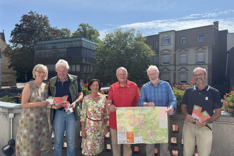 Foto: Denise Rüscher/Stadt Menden Sechs Personen stehen bei sonnigem Wetter auf einer Terrasse und präsentieren Wanderkarten sowie Informationsbroschüren. Zwei Männer halten eine großformatige Karte der Umgebung, während die anderen Prospekte in den Händen haben. Im Hintergrund sind Bäume, Blumenkästen und Gebäude der Innenstadt zu sehen.
