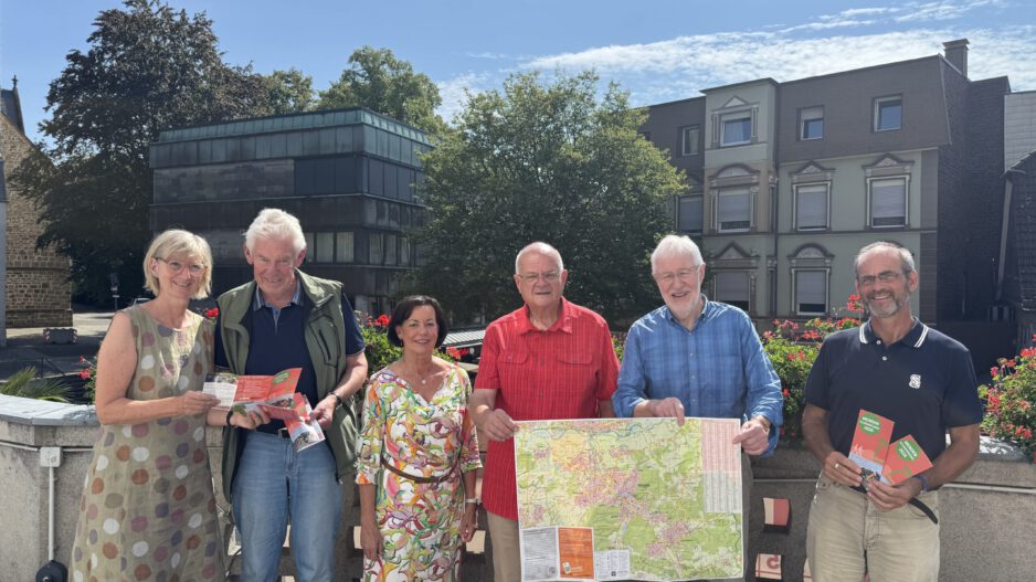 Sechs Personen stehen bei sonnigem Wetter auf einer Terrasse und präsentieren Wanderkarten sowie Informationsbroschüren. Zwei Männer halten eine großformatige Karte der Umgebung, während die anderen Prospekte in den Händen haben. Im Hintergrund sind Bäume, Blumenkästen und Gebäude der Innenstadt zu sehen.