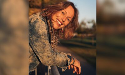 Eine lächelnde Frau mit schulterlangen braunen Haaren lehnt sich im warmen Abendlicht über ein Geländer. Sie trägt eine Jeansjacke; im unscharfen Hintergrund sind herbstlich gefärbte Bäume zu sehen.