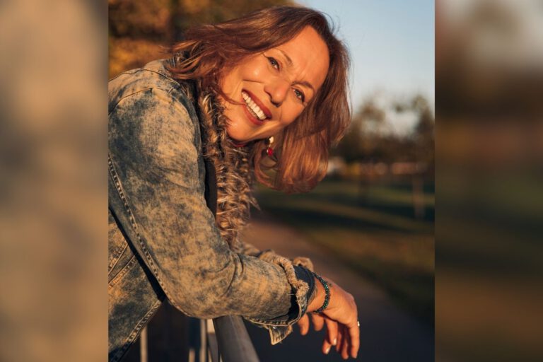 Eine lächelnde Frau mit schulterlangen braunen Haaren lehnt sich im warmen Abendlicht über ein Geländer. Sie trägt eine Jeansjacke; im unscharfen Hintergrund sind herbstlich gefärbte Bäume zu sehen.