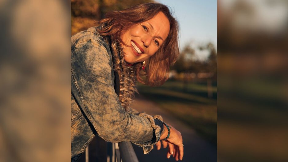 Eine lächelnde Frau mit schulterlangen braunen Haaren lehnt sich im warmen Abendlicht über ein Geländer. Sie trägt eine Jeansjacke; im unscharfen Hintergrund sind herbstlich gefärbte Bäume zu sehen.