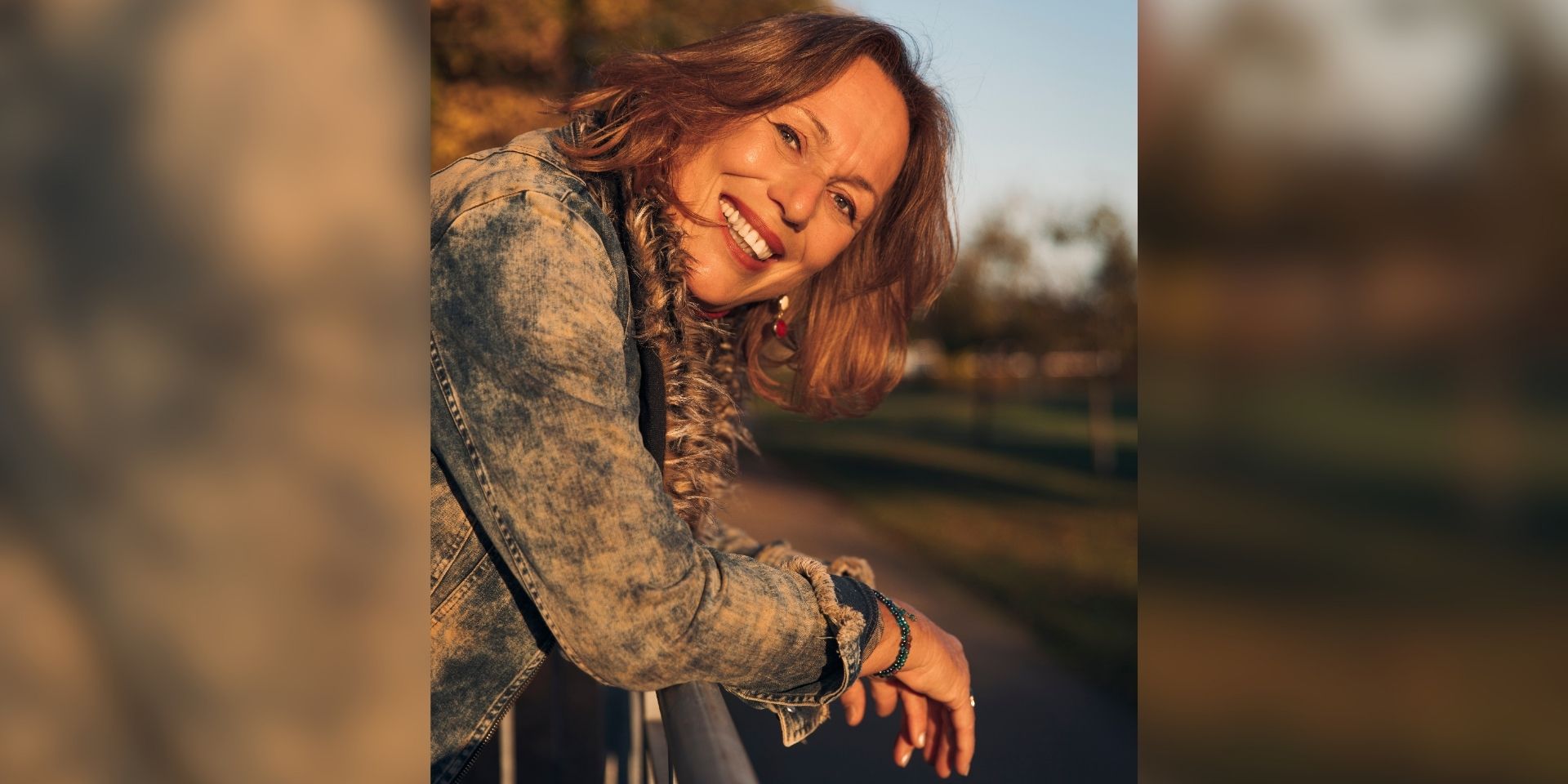Eine lächelnde Frau mit schulterlangen braunen Haaren lehnt sich im warmen Abendlicht über ein Geländer. Sie trägt eine Jeansjacke; im unscharfen Hintergrund sind herbstlich gefärbte Bäume zu sehen.