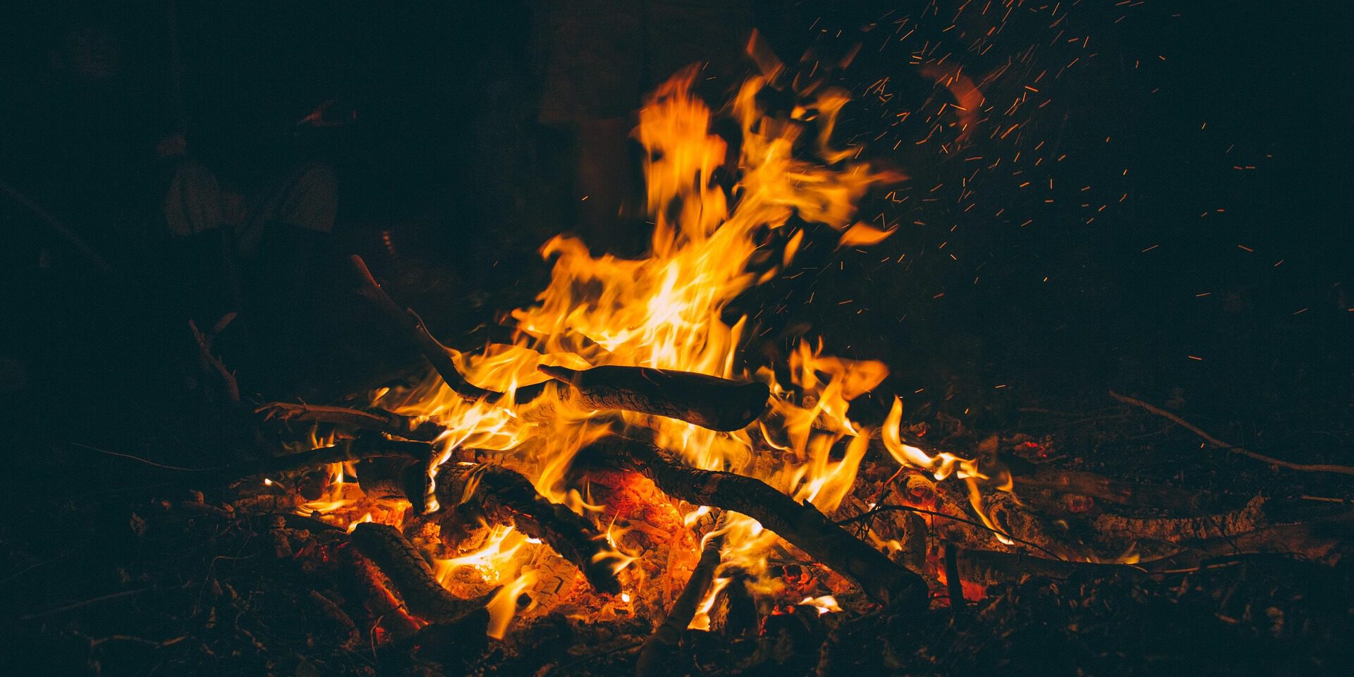 Ein kräftiges Lagerfeuer brennt in der Dunkelheit. Orangefarbene Flammen lodern zwischen Ästen und Holzscheiten, während zahlreiche Funken in die Luft steigen. Im schwach beleuchteten Hintergrund sind schemenhaft Personen rund um das Feuer zu erkennen.