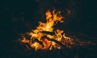 Ein kräftiges Lagerfeuer brennt in der Dunkelheit. Orangefarbene Flammen lodern zwischen Ästen und Holzscheiten, während zahlreiche Funken in die Luft steigen. Im schwach beleuchteten Hintergrund sind schemenhaft Personen rund um das Feuer zu erkennen.