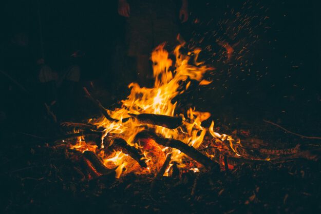 Ein kräftiges Lagerfeuer brennt in der Dunkelheit. Orangefarbene Flammen lodern zwischen Ästen und Holzscheiten, während zahlreiche Funken in die Luft steigen. Im schwach beleuchteten Hintergrund sind schemenhaft Personen rund um das Feuer zu erkennen.