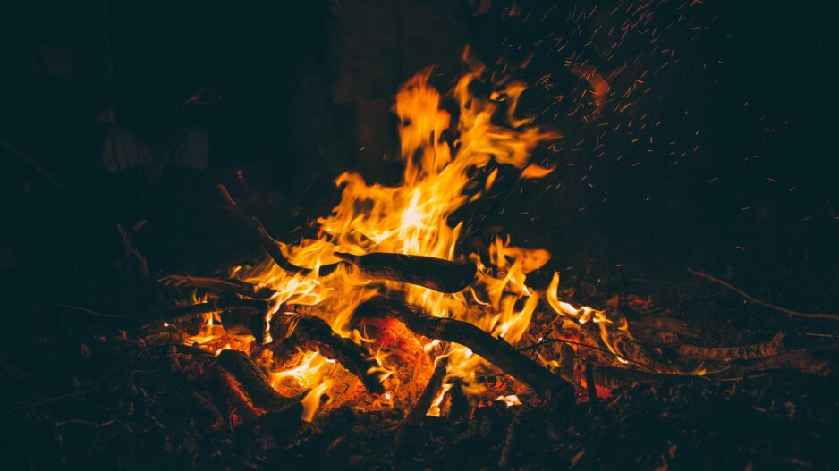 Ein kräftiges Lagerfeuer brennt in der Dunkelheit. Orangefarbene Flammen lodern zwischen Ästen und Holzscheiten, während zahlreiche Funken in die Luft steigen. Im schwach beleuchteten Hintergrund sind schemenhaft Personen rund um das Feuer zu erkennen.