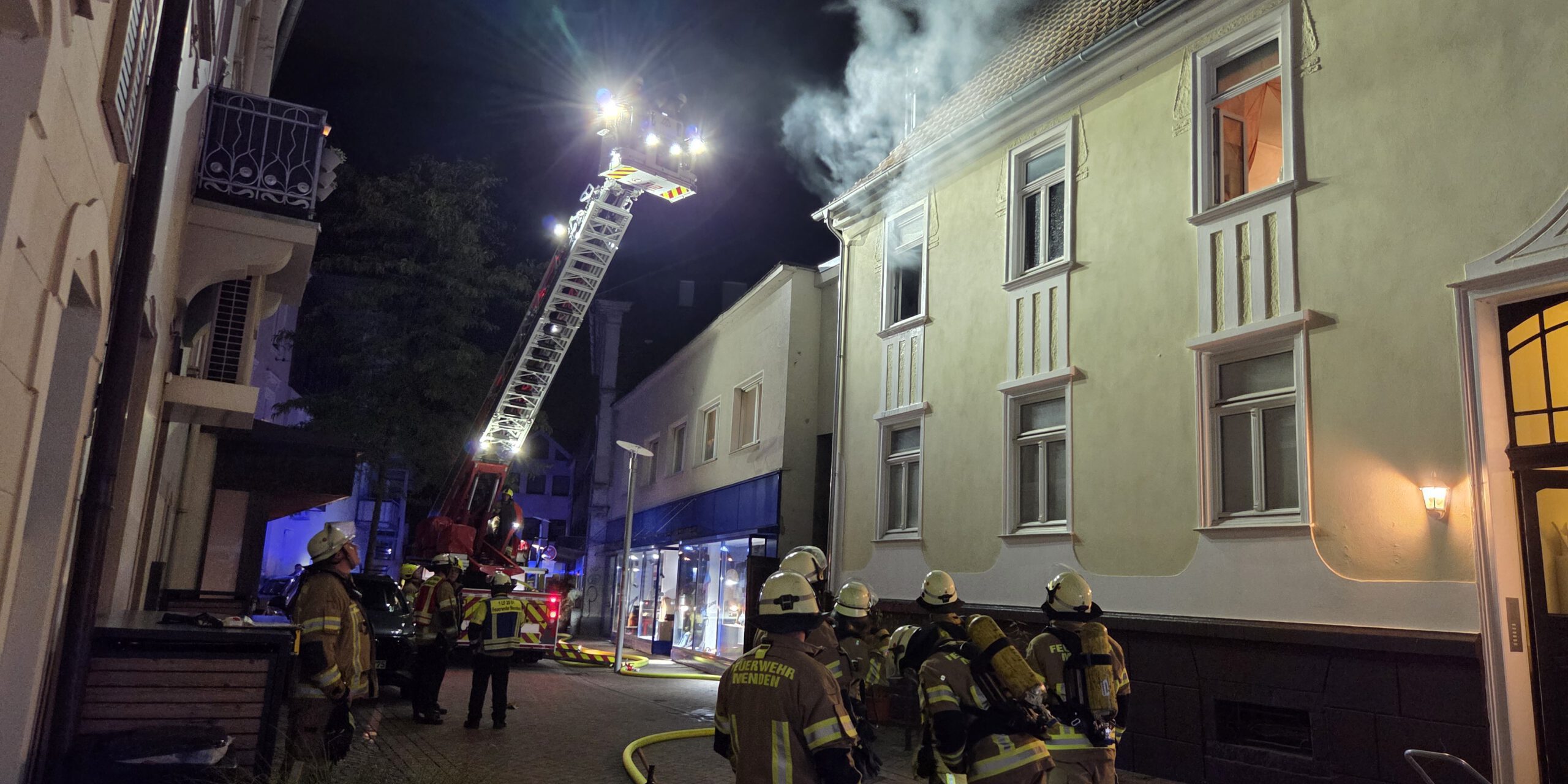 Nächtlicher Feuerwehreinsatz in einer Innenstadtstraße: Mehrere Einsatzkräfte stehen vor einem mehrstöckigen Gebäude, aus dessen Dachbereich Rauch aufsteigt. Eine ausgefahrene Drehleiter ist auf das Dach gerichtet; Schläuche und weitere Einsatzgeräte liegen auf der Straße.