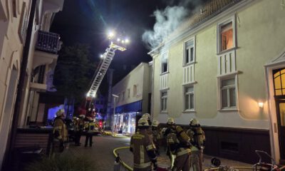 Nächtlicher Feuerwehreinsatz in einer Innenstadtstraße: Mehrere Einsatzkräfte stehen vor einem mehrstöckigen Gebäude, aus dessen Dachbereich Rauch aufsteigt. Eine ausgefahrene Drehleiter ist auf das Dach gerichtet; Schläuche und weitere Einsatzgeräte liegen auf der Straße.