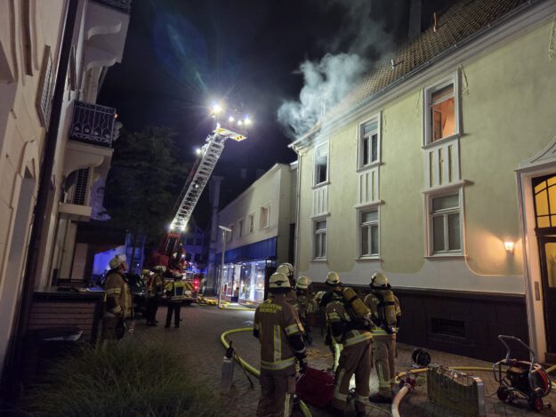 Nächtlicher Feuerwehreinsatz in einer Innenstadtstraße: Mehrere Einsatzkräfte stehen vor einem mehrstöckigen Gebäude, aus dessen Dachbereich Rauch aufsteigt. Eine ausgefahrene Drehleiter ist auf das Dach gerichtet; Schläuche und weitere Einsatzgeräte liegen auf der Straße.