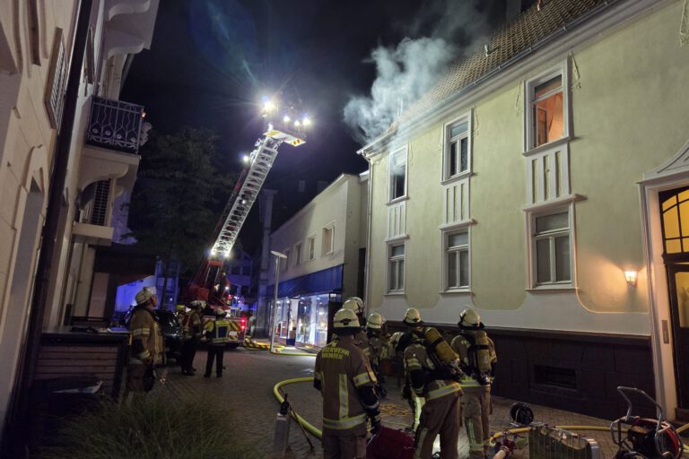 Nächtlicher Feuerwehreinsatz in einer Innenstadtstraße: Mehrere Einsatzkräfte stehen vor einem mehrstöckigen Gebäude, aus dessen Dachbereich Rauch aufsteigt. Eine ausgefahrene Drehleiter ist auf das Dach gerichtet; Schläuche und weitere Einsatzgeräte liegen auf der Straße.