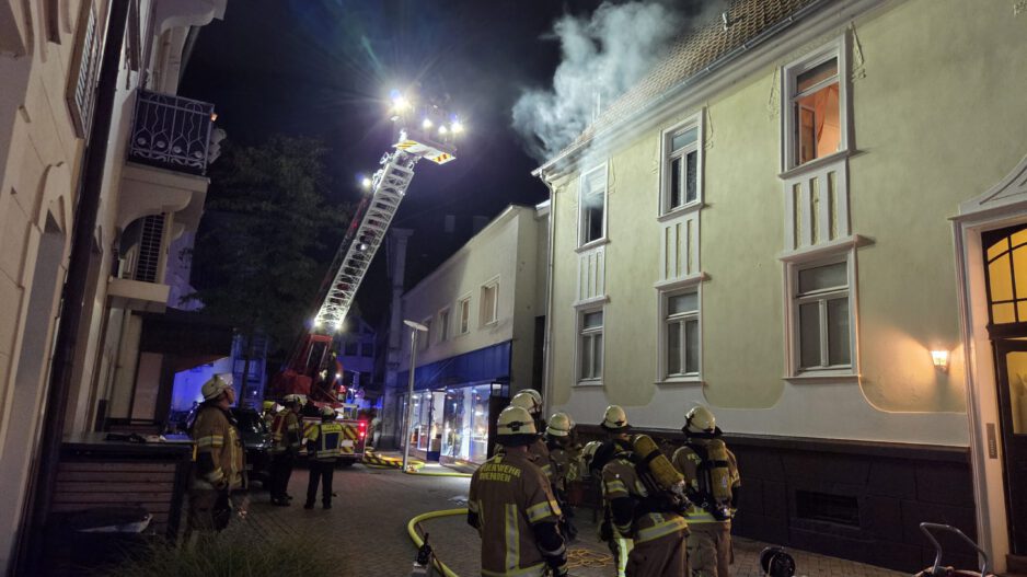 Nächtlicher Feuerwehreinsatz in einer Innenstadtstraße: Mehrere Einsatzkräfte stehen vor einem mehrstöckigen Gebäude, aus dessen Dachbereich Rauch aufsteigt. Eine ausgefahrene Drehleiter ist auf das Dach gerichtet; Schläuche und weitere Einsatzgeräte liegen auf der Straße.