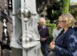 Drei Personen betrachten bei sonnigem Wetter eine große graue Metallskulptur mit spiegelnden Kugeln. Sie stehen in einem begrünten Außenbereich; zwei von ihnen halten Gläser in den Händen.
