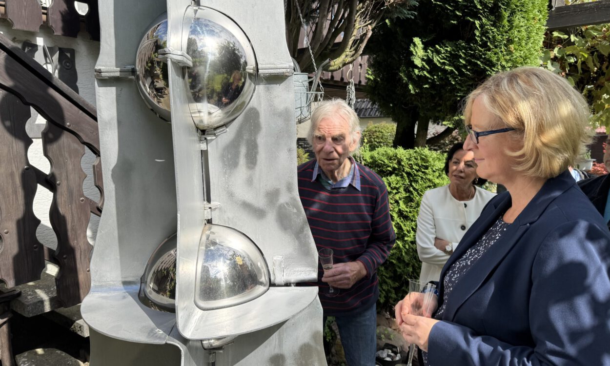 Drei Personen betrachten bei sonnigem Wetter eine große graue Metallskulptur mit spiegelnden Kugeln. Sie stehen in einem begrünten Außenbereich; zwei von ihnen halten Gläser in den Händen.