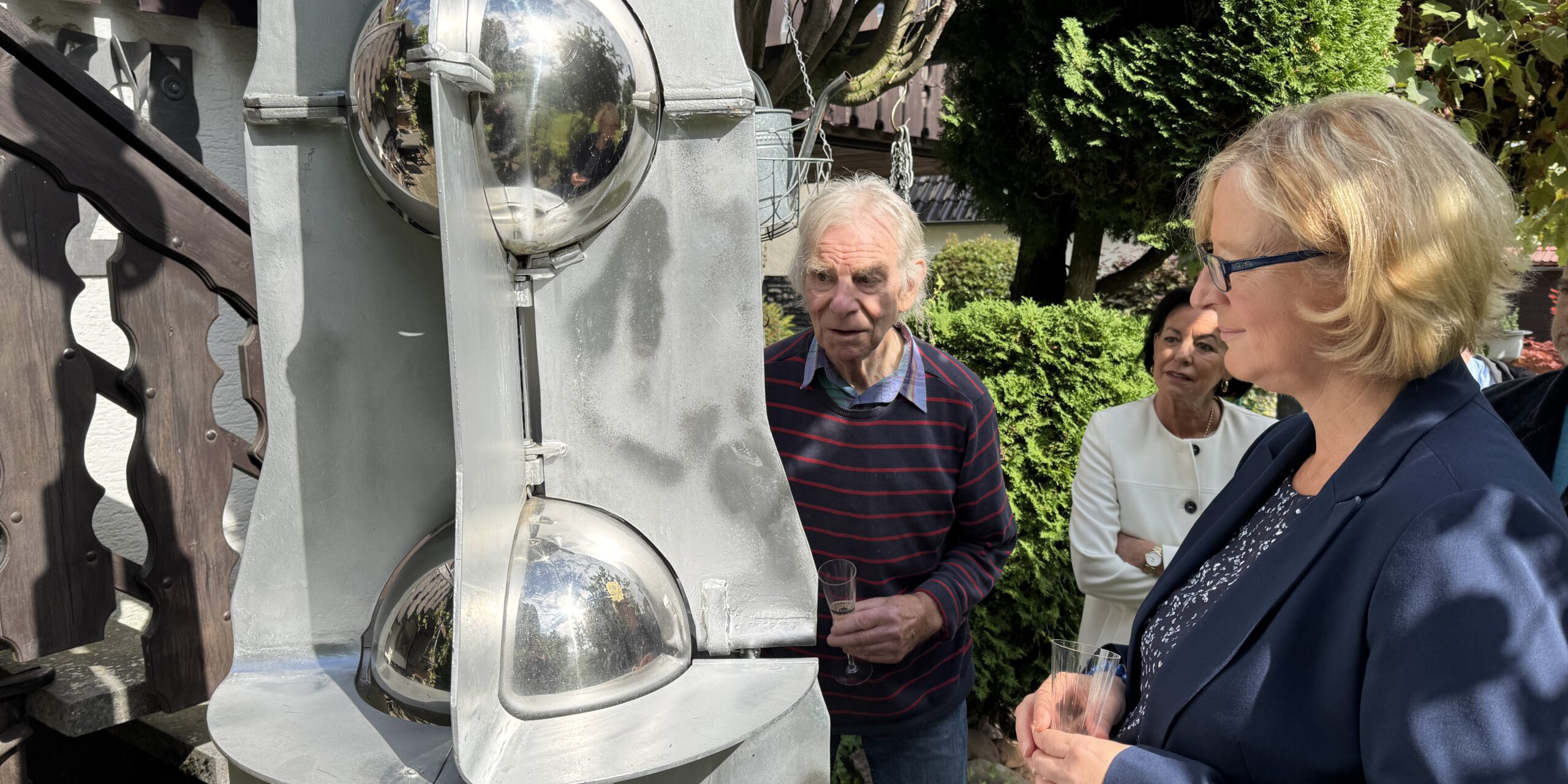 Drei Personen betrachten bei sonnigem Wetter eine große graue Metallskulptur mit spiegelnden Kugeln. Sie stehen in einem begrünten Außenbereich; zwei von ihnen halten Gläser in den Händen.