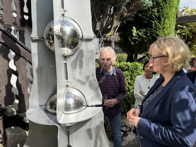 Foto: D. Rüscher/Stadt Menden Drei Personen betrachten bei sonnigem Wetter eine große graue Metallskulptur mit spiegelnden Kugeln. Sie stehen in einem begrünten Außenbereich; zwei von ihnen halten Gläser in den Händen.