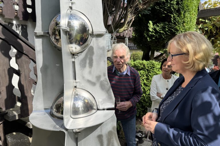 Drei Personen betrachten bei sonnigem Wetter eine große graue Metallskulptur mit spiegelnden Kugeln. Sie stehen in einem begrünten Außenbereich; zwei von ihnen halten Gläser in den Händen.