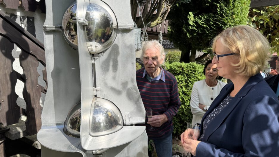 Drei Personen betrachten bei sonnigem Wetter eine große graue Metallskulptur mit spiegelnden Kugeln. Sie stehen in einem begrünten Außenbereich; zwei von ihnen halten Gläser in den Händen.