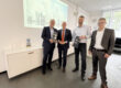 Vier Männer stehen in einem Besprechungsraum vor einer Präsentation mit der Aufschrift „Die Welt von ECO Schulte“. Zwei von ihnen halten schwarze Präsentationsboxen in den Händen.