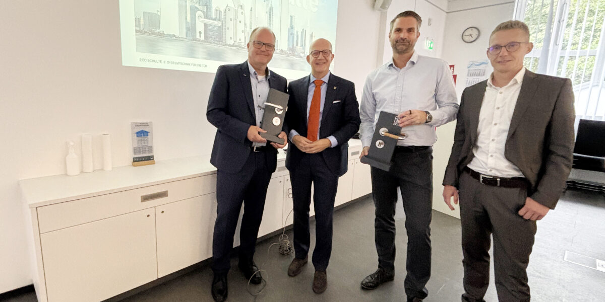 Vier Männer stehen in einem Besprechungsraum vor einer Präsentation mit der Aufschrift „Die Welt von ECO Schulte“. Zwei von ihnen halten schwarze Präsentationsboxen in den Händen.